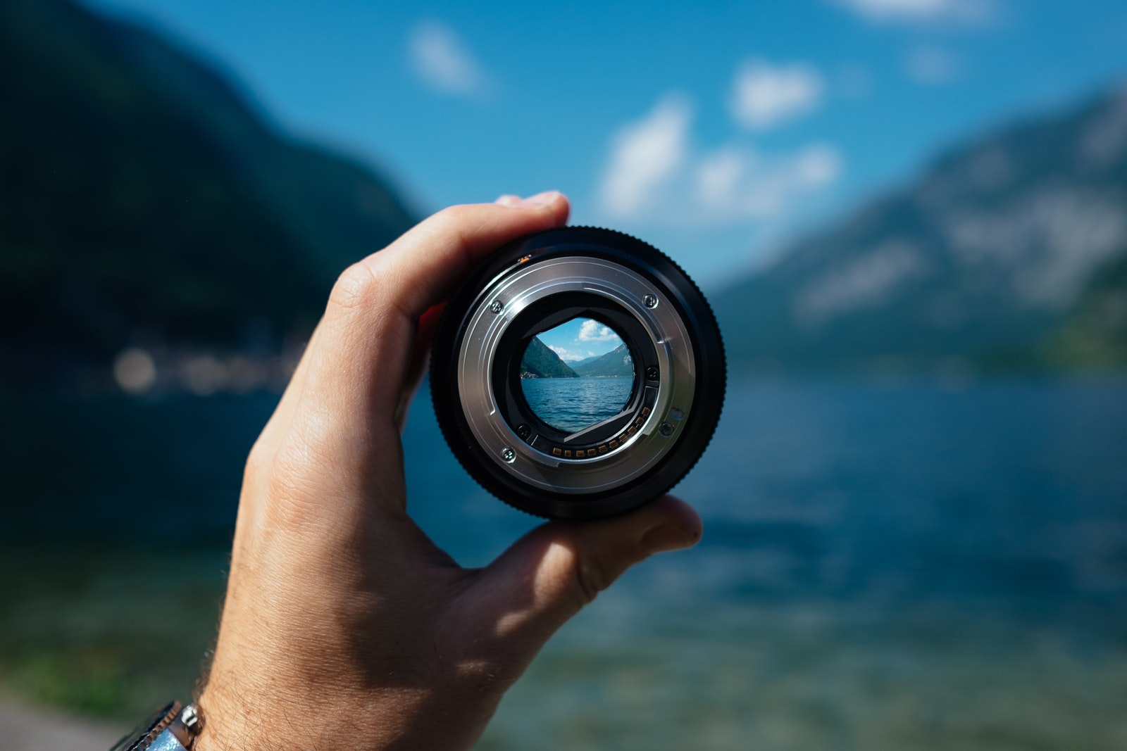 A camera lens in a hand with a view behind of mountains and a body of water